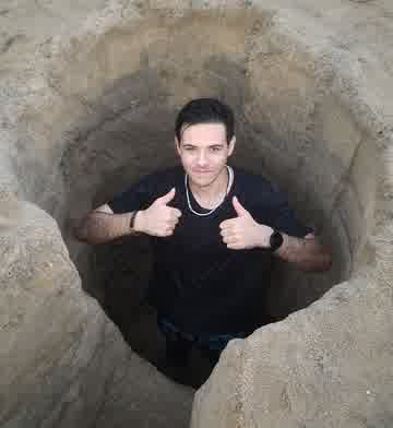 A man standing chest-deep in a hole dug in beach sand, giving two thumbs up
